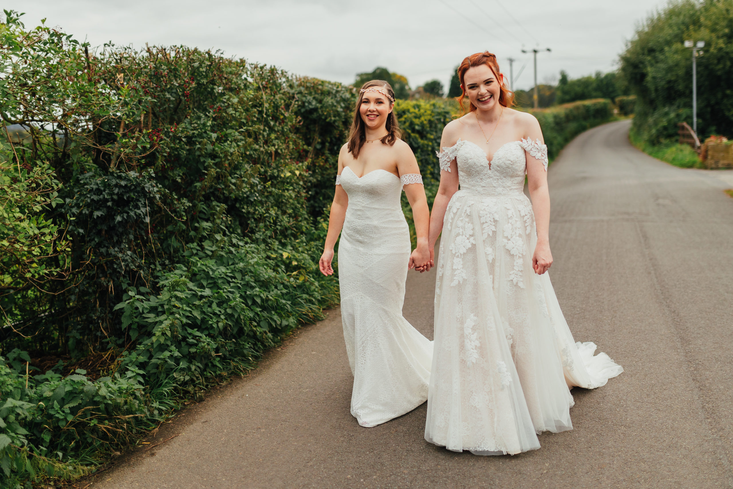a lesbian couple in white wedding dresses smiling and holding hands walking towards the camera
