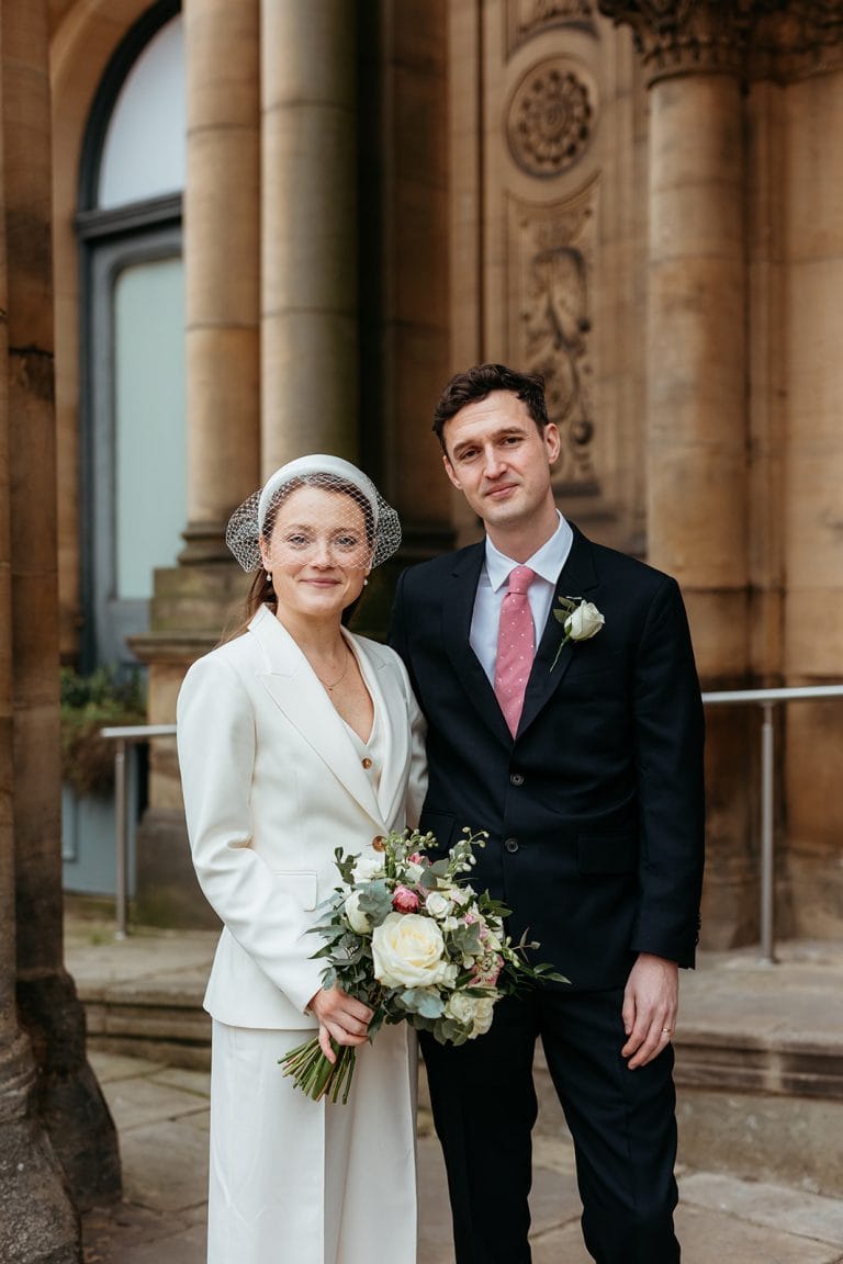 Bride and groom in wedding attire outside historic building in Manchester.