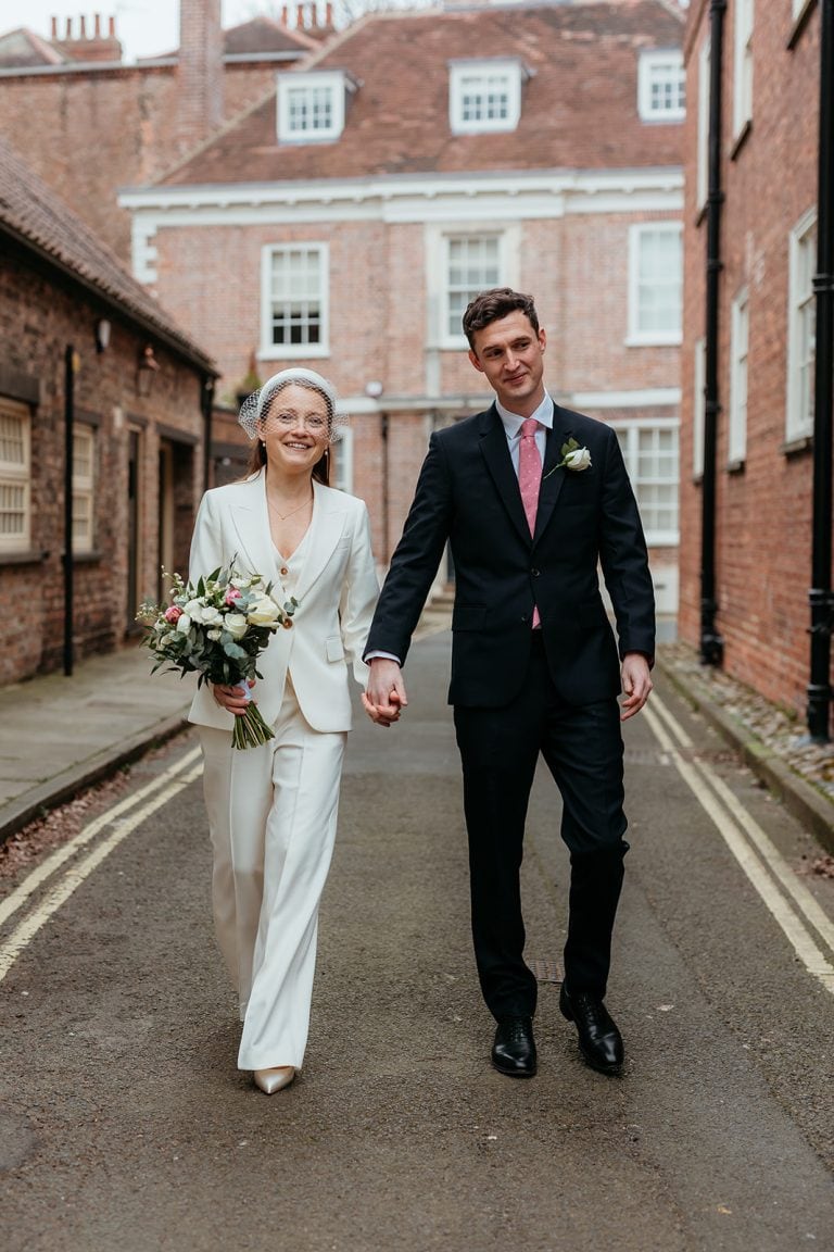Happy wedding couple walking hand in hand in city street with historic brick buildings.